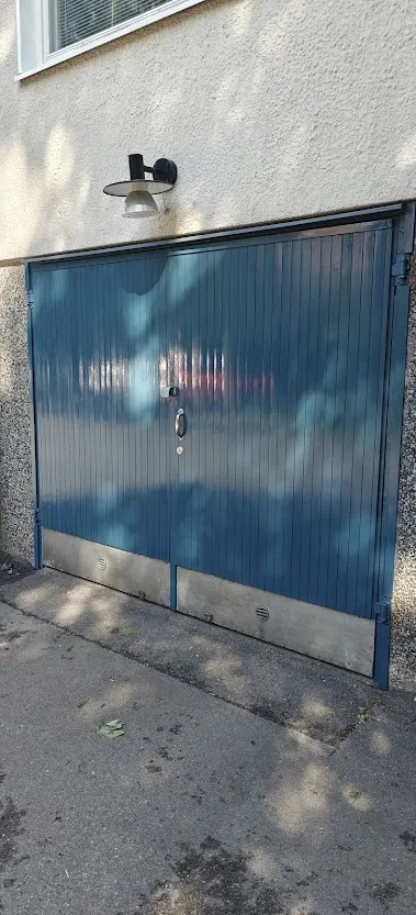 Sininen pystypaneelinen autotallin ovi, jossa on kahva ja lukko, ja sen yläpuolella on seinävalaisin.